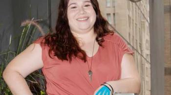 A young woman stands in an urban environment. She is smiling and appears confident. She has blue therapy tape on the back of each hand. The tape strips run from each fingertip to her wrist.